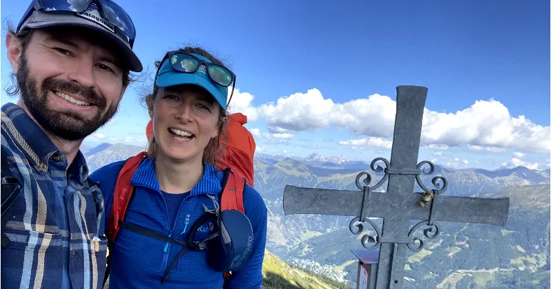Twee wandelaars glimlachen naar de camera op een bergtop naast een metalen kruis, met op de achtergrond een blauwe lucht, wolken en bergtoppen in de verte.